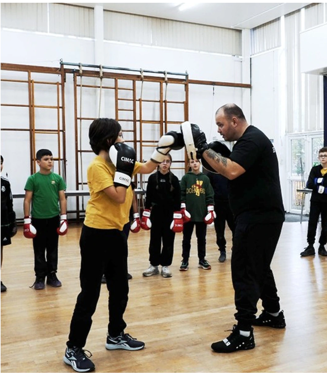 A group of schoolchildren in a gymnasium watch as a boxing coach demonstrates a punching technique with a student wearing gloves.